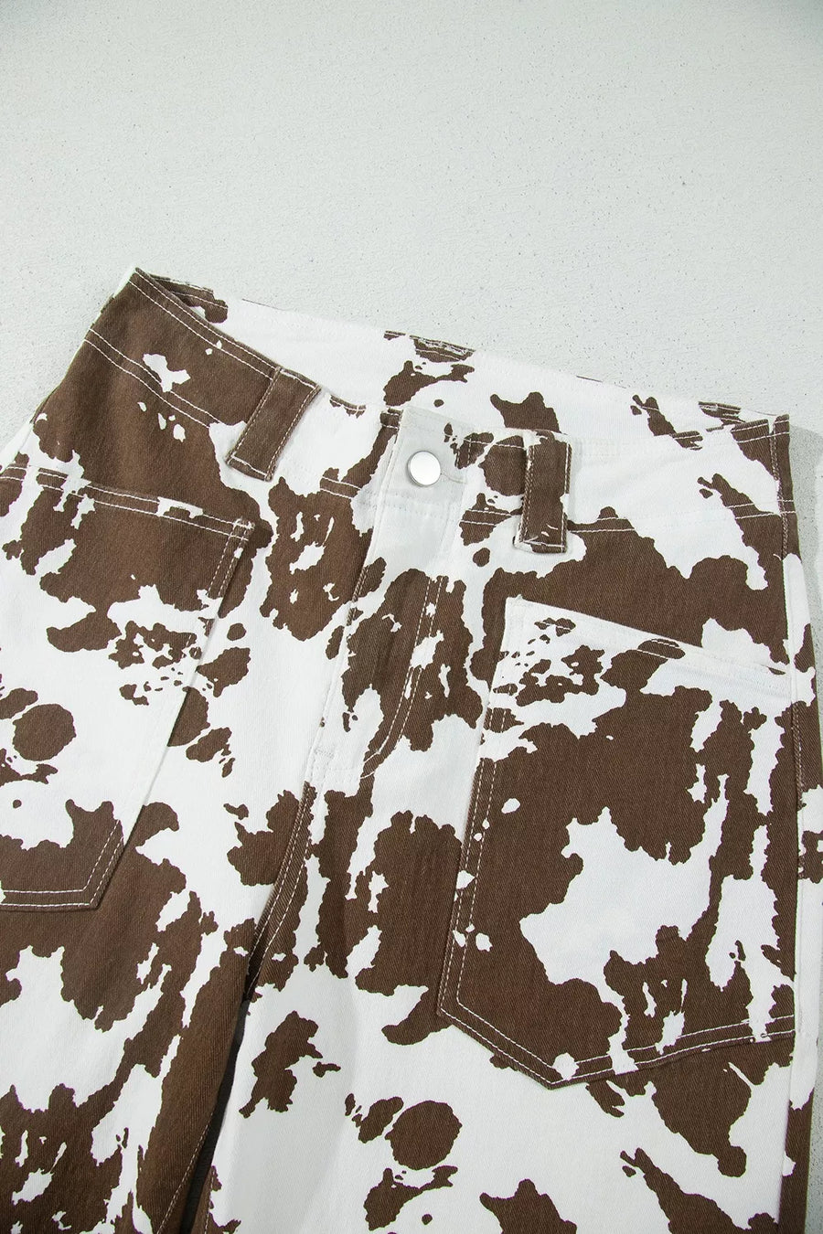 Close-up of a pair of pants with a brown and white abstract splatter pattern, showing the waistband, belt loops, button, and front pockets.