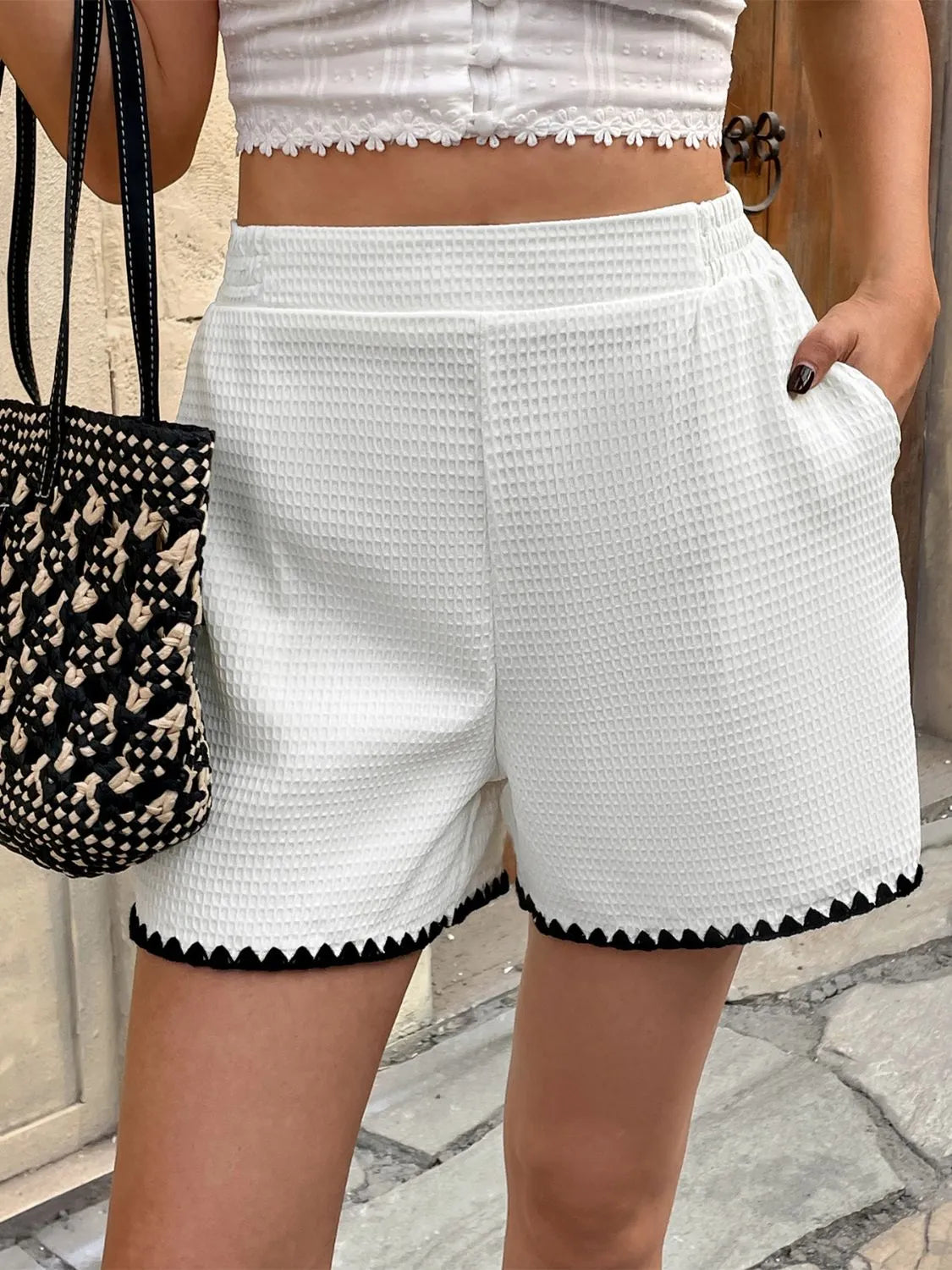 Person wearing white shorts with black trim, holding a woven bag on a stone pavement.