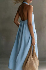 Woman wearing a light blue dress with an open back, holding a woven bag against a neutral background.