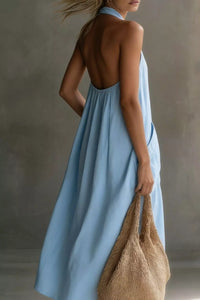 Woman wearing a light blue dress with an open back, holding a woven bag against a neutral background.