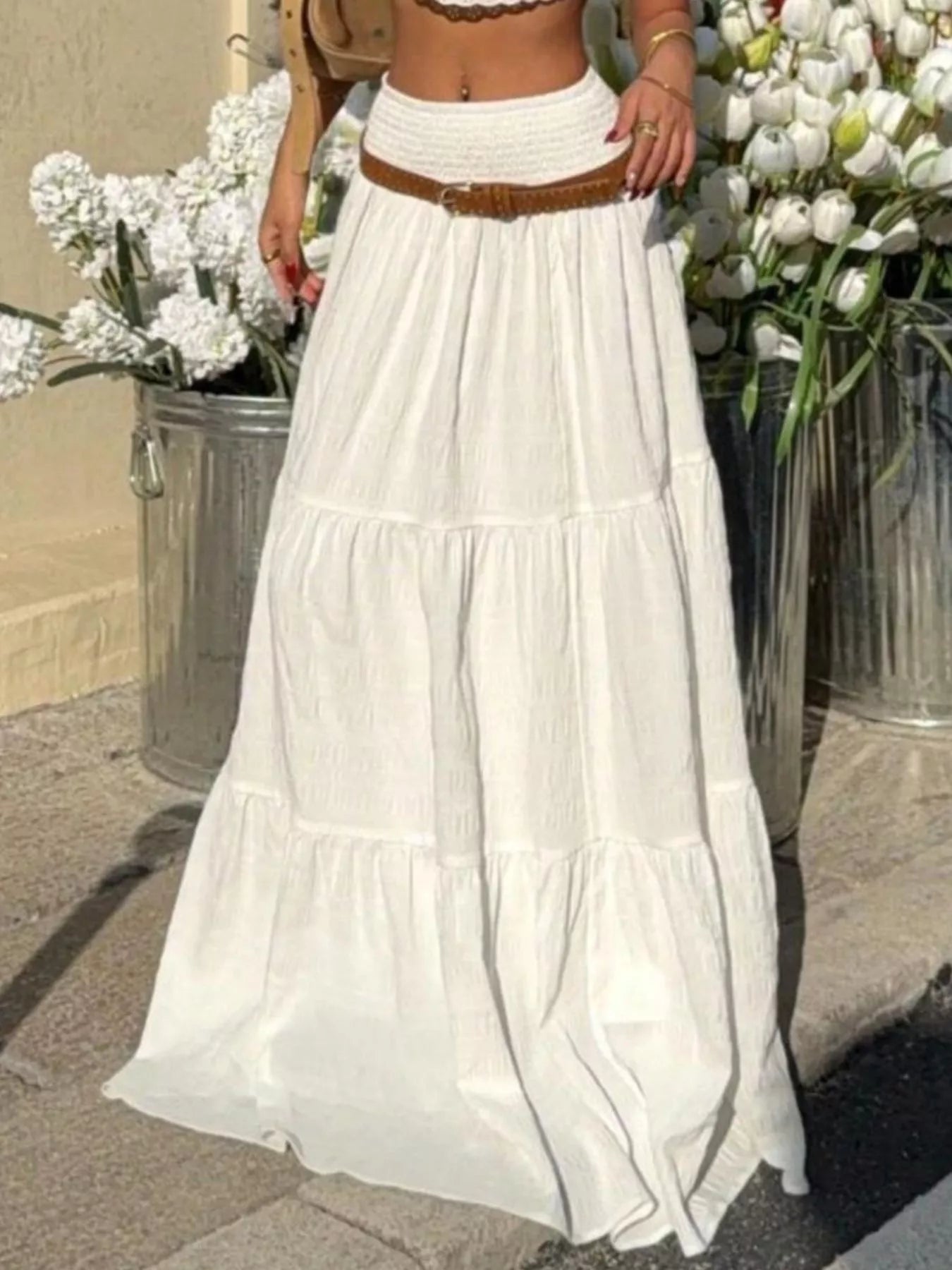 Person wearing a white tiered skirt with a brown belt, standing in front of floral arrangements.