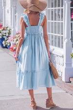 Woman in a light blue dress with ruffled straps and hem, wearing a wide-brimmed hat, standing on a sidewalk with flowers and a white railing in the background.