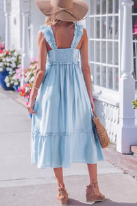Woman in a light blue dress with ruffled straps and hem, wearing a wide-brimmed hat, standing on a sidewalk with flowers and a white railing in the background.