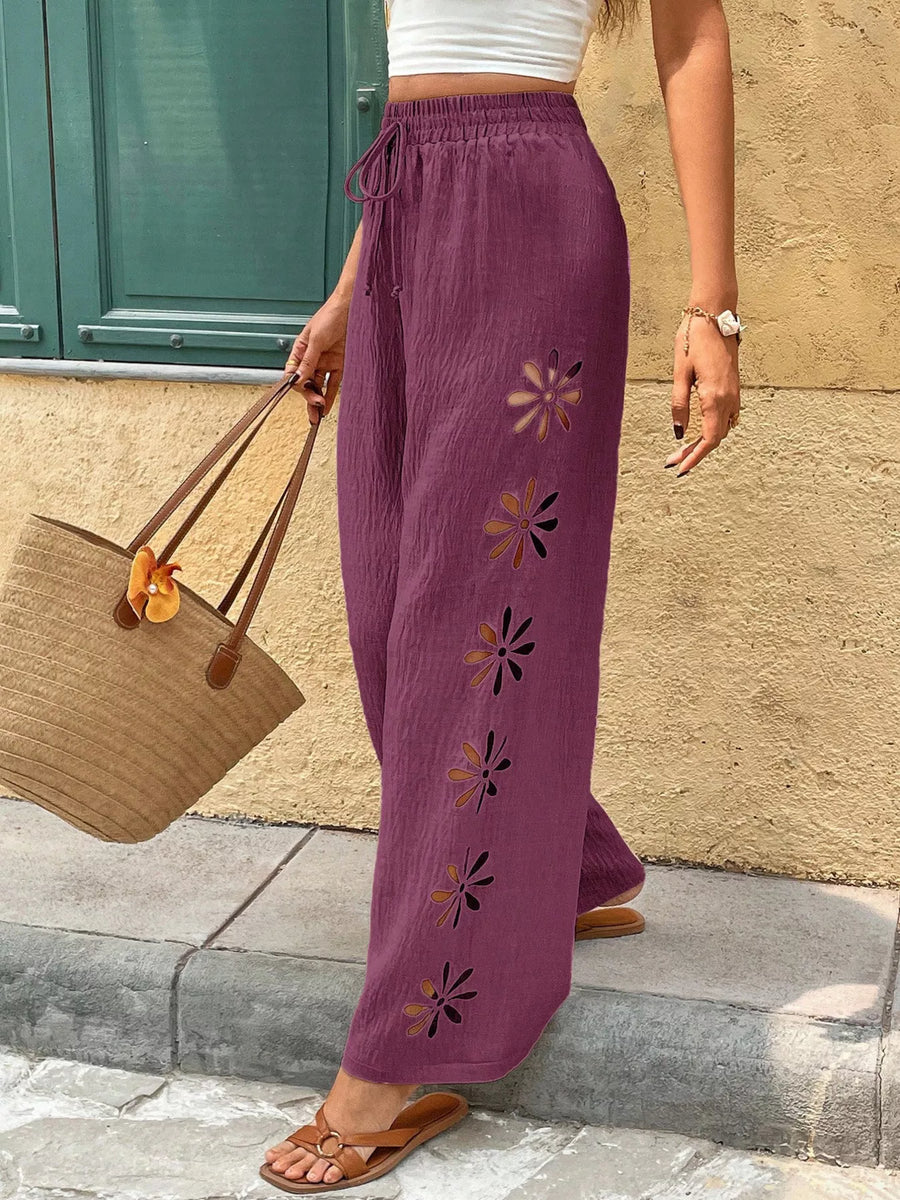 Person wearing purple pants with floral embroidery, standing against a stone wall.