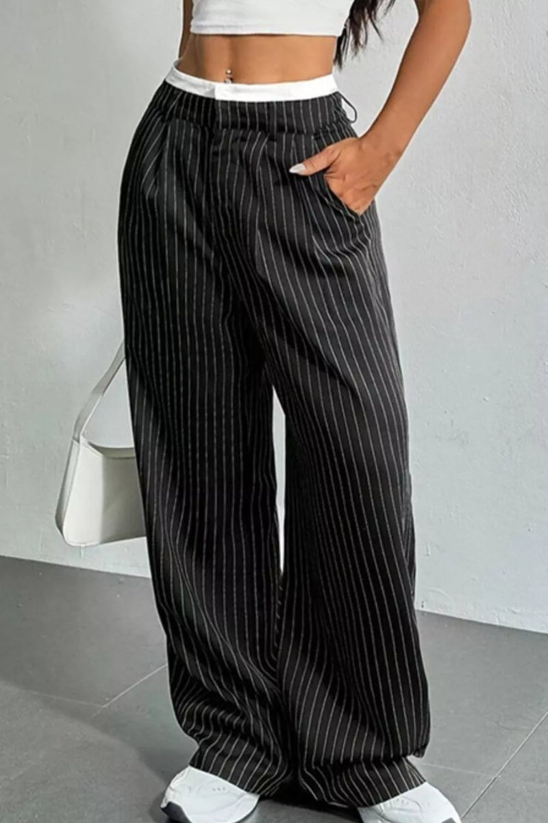A person wearing loose black wide-leg pants with thin white vertical pinstripes, paired with a white crop top and white sneakers, standing against a light gray wall.