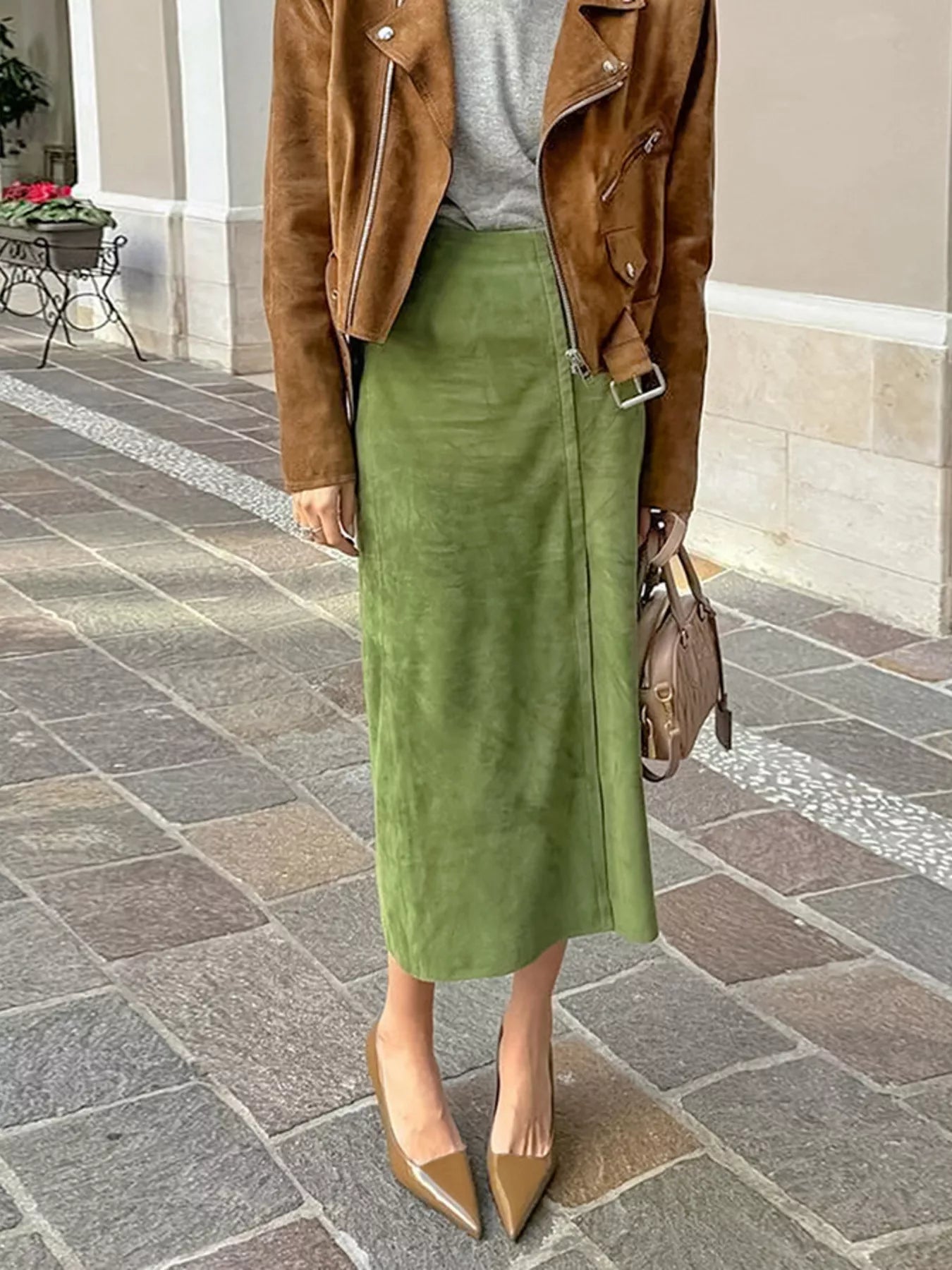 Person wearing a brown jacket, green skirt, and beige shoes on a stone pavement.