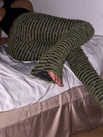 A person reclining on a bed wearing chunky knit striped lounge pants in olive green and black, with a black top partially visible.