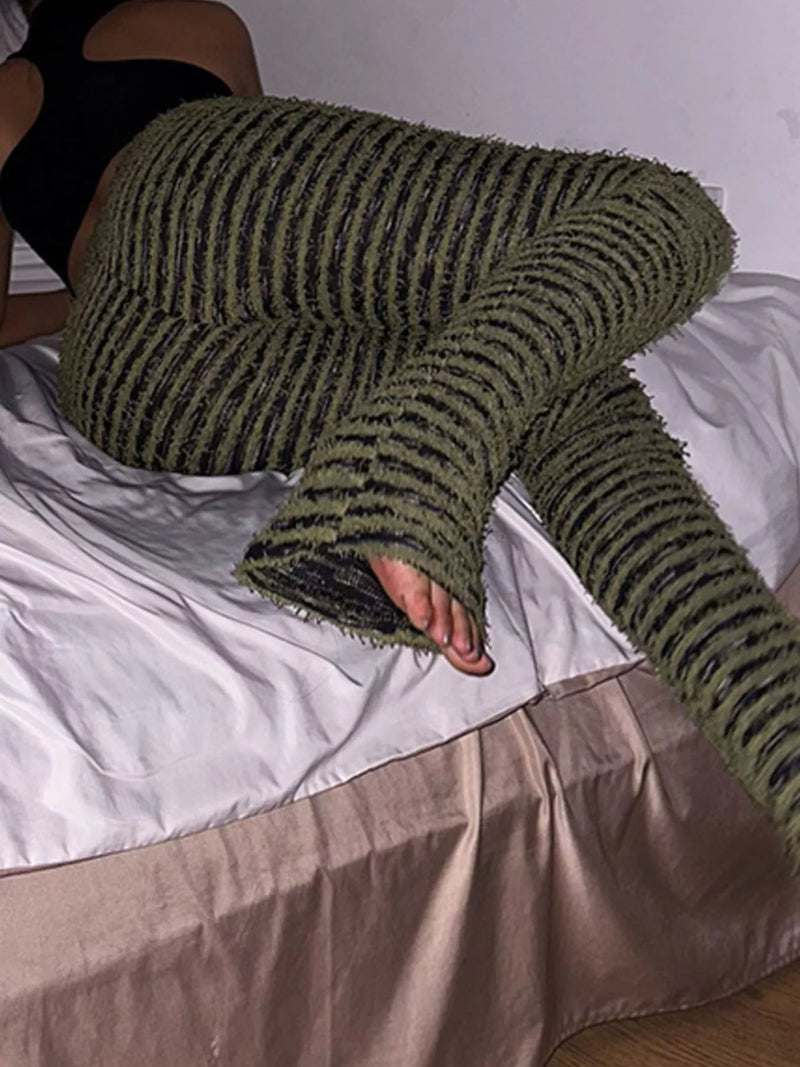 A person reclining on a bed wearing chunky knit striped lounge pants in olive green and black, with a black top partially visible.