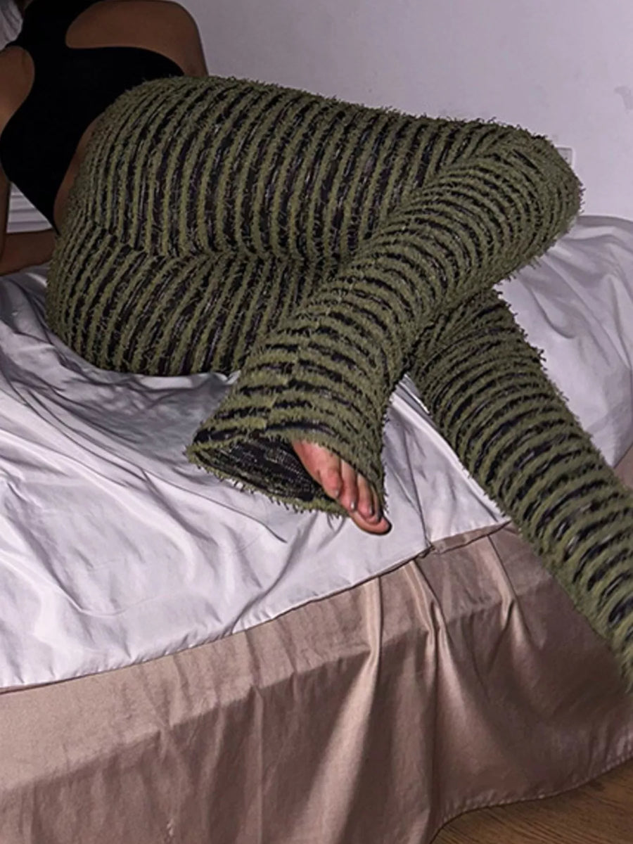 A person reclining on a bed wearing chunky knit striped lounge pants in olive green and black, with a black top partially visible.