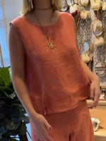 Person wearing a sleeveless orange top with a sun pendant necklace in a store setting.