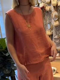 Person wearing a sleeveless orange top with a sun pendant necklace in a store setting.