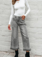 Person wearing gray wide-leg jeans and a white long-sleeve top against a light gray wall.