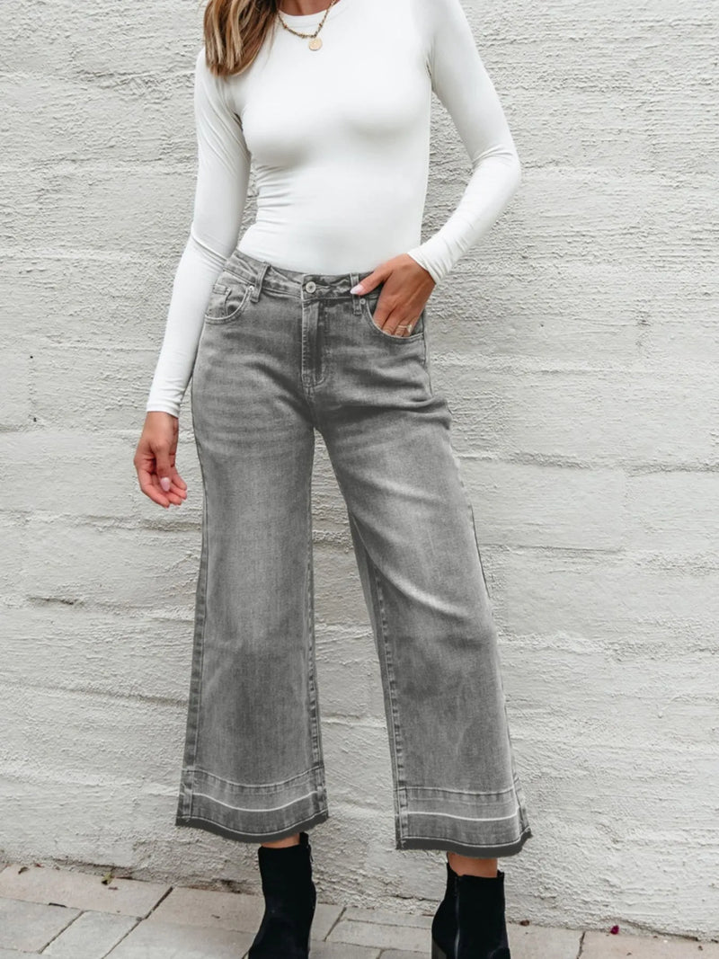 Person wearing gray wide-leg jeans and a white long-sleeve top against a light gray wall.