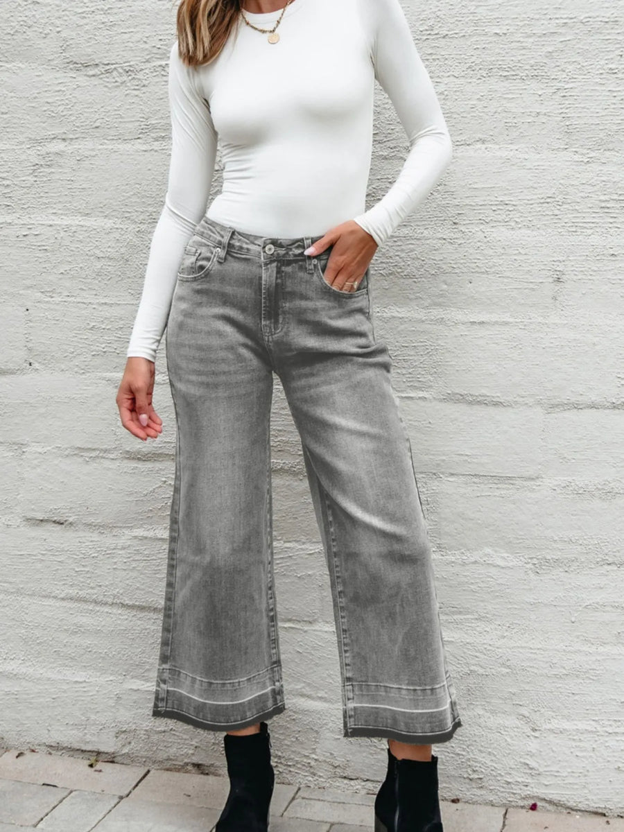 Person wearing gray wide-leg jeans and a white long-sleeve top against a light gray wall.