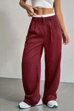 A person wearing loose, wide-leg trousers in deep burgundy with thin white vertical pinstripes, paired with a white crop top and white sneakers, standing against a light gray wall.