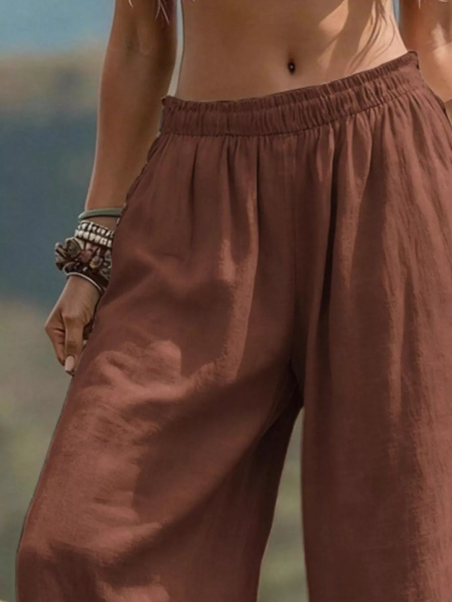 A close-up of a person wearing loose, wide-leg brown pants with an elastic waistband, shown outdoors against a blurred natural background.