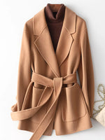 A camel-colored belted coat with wide lapels and front patch pockets, styled over a dark brown ribbed turtleneck sweater. A small cream handbag with a gold chain is partially visible on the right side against a plain white background.