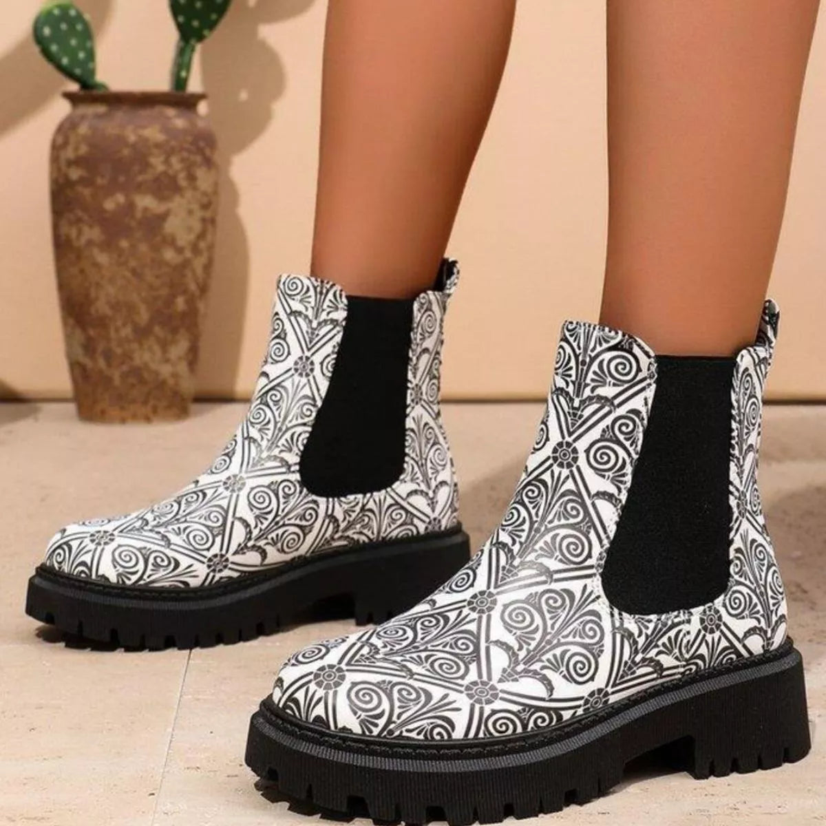 Black and white patterned boots with black soles worn by a person, with a vase and plant in the background.