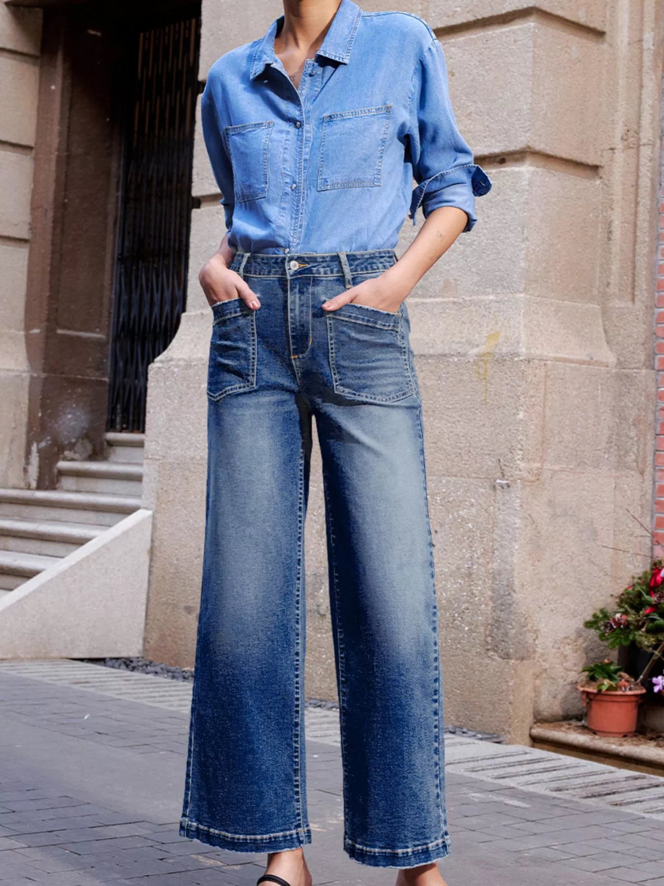 Person wearing a blue denim shirt and wide-leg jeans standing against a stone building.