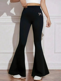 Black flared pants with a pink bow detail worn by a person on a light wooden floor.