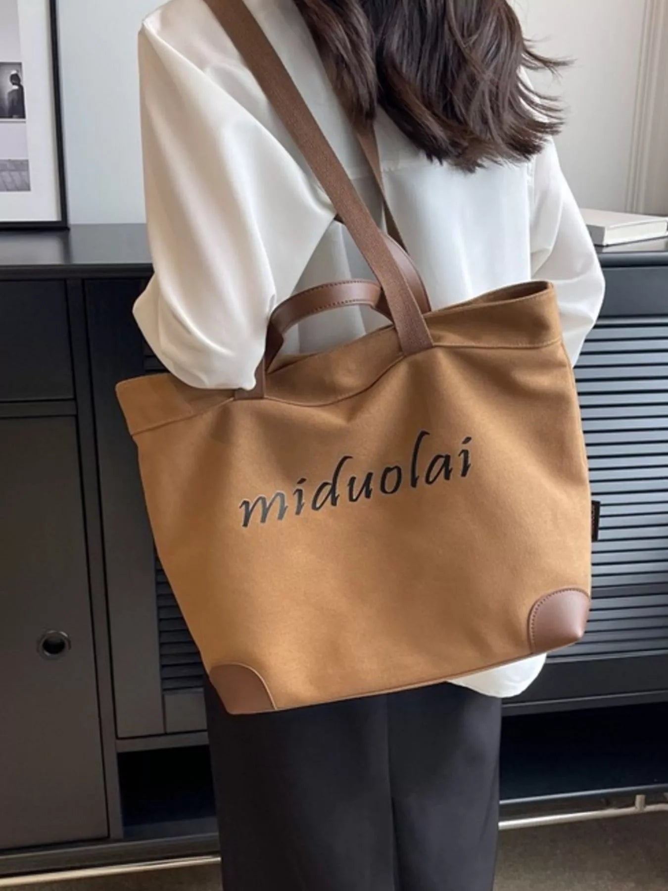 Person holding a brown tote bag with 'miduolai' branding indoors.