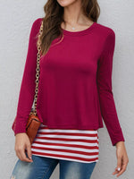 A woman wearing a long-sleeve crewneck top in deep magenta, paired with a red and white striped tunic and blue jeans, carrying a small brown crossbody bag with a gold chain strap against a textured gray wall.