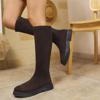 Person wearing black knee-high boots in a room with a wooden chair and beige tiles.