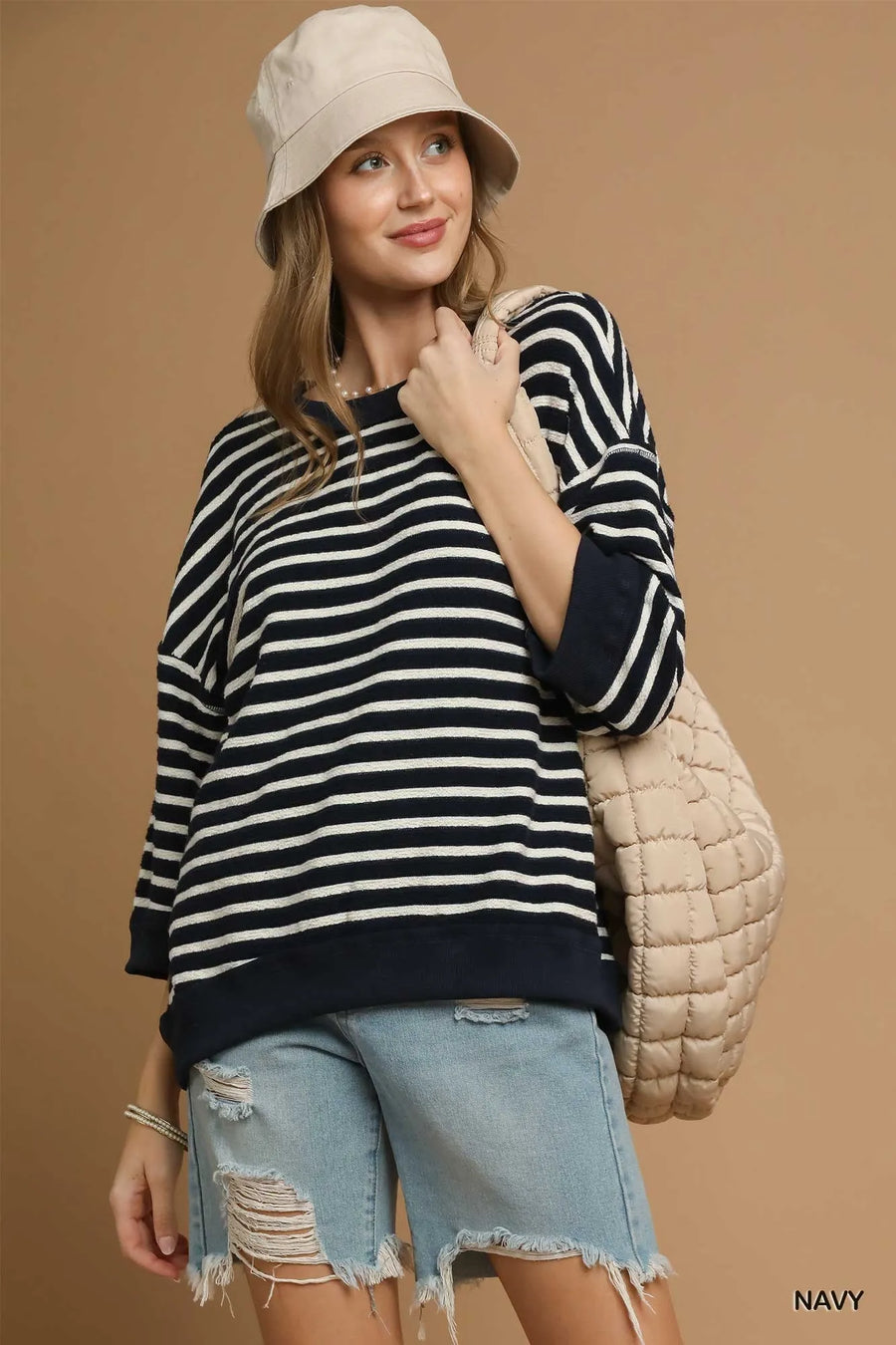 Woman wearing a striped sweater, denim shorts, and a beige bucket hat against a brown background.