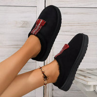 Black slip-on shoes with red and white patterned straps worn by a person on a light wooden floor.