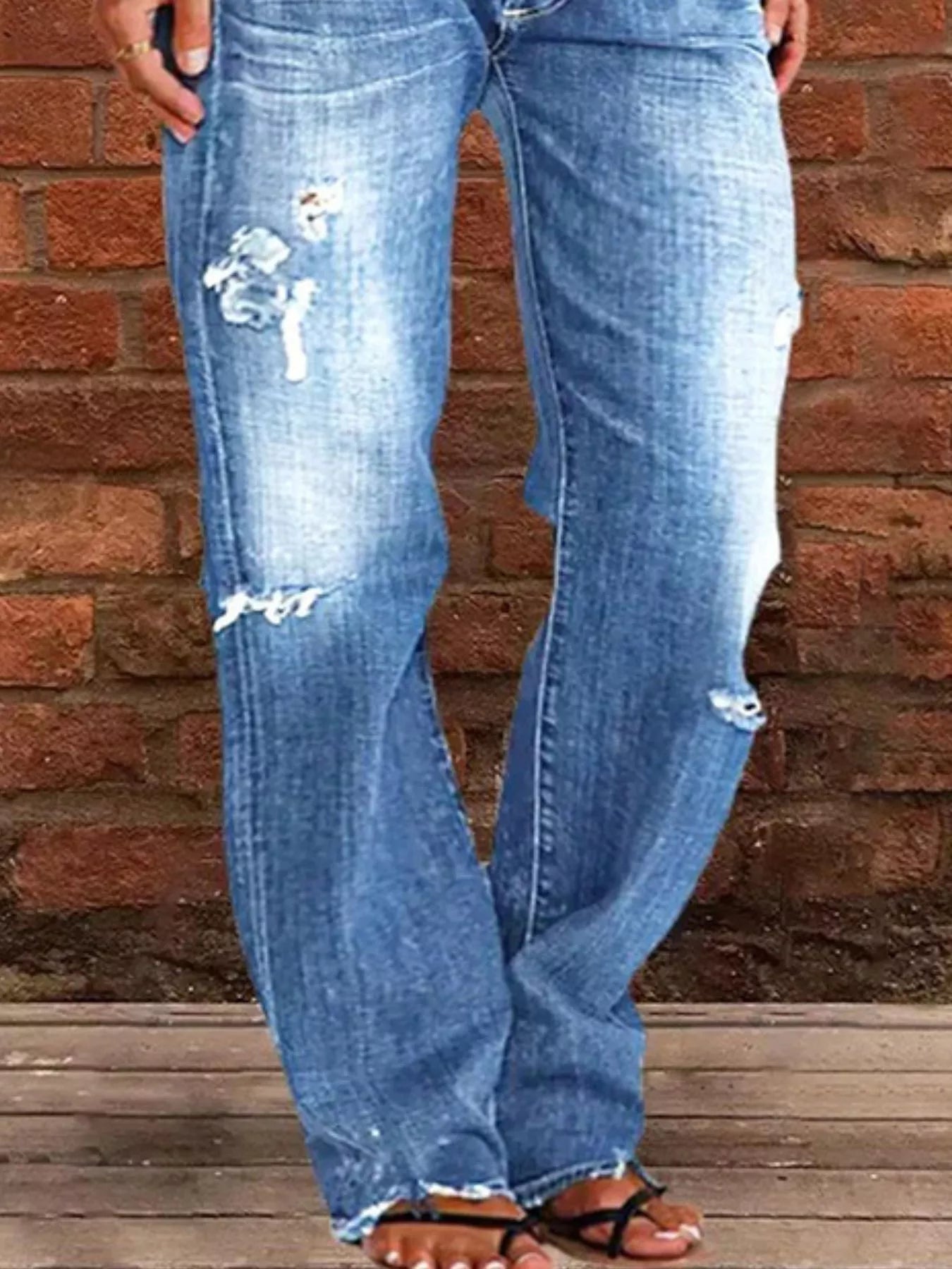 Blue jeans with distressing details worn by a person against a brick wall.