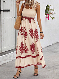 Woman wearing a beige dress with red patterns, standing against a white wall.