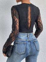 Person wearing a black lace long-sleeve top and blue jeans against a white wall.