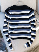 A long-sleeve ribbed knit sweater with horizontal stripes in navy blue, slate blue, and white, displayed on a clear hanger over a light beige upholstered chair.