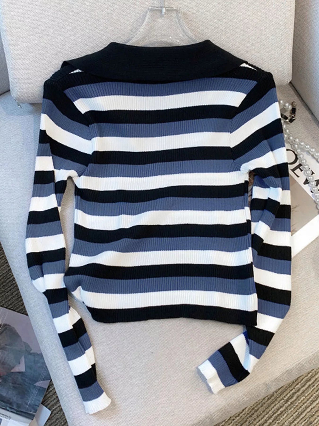 A long-sleeve ribbed knit sweater with horizontal stripes in navy blue, slate blue, and white, displayed on a clear hanger over a light beige upholstered chair.
