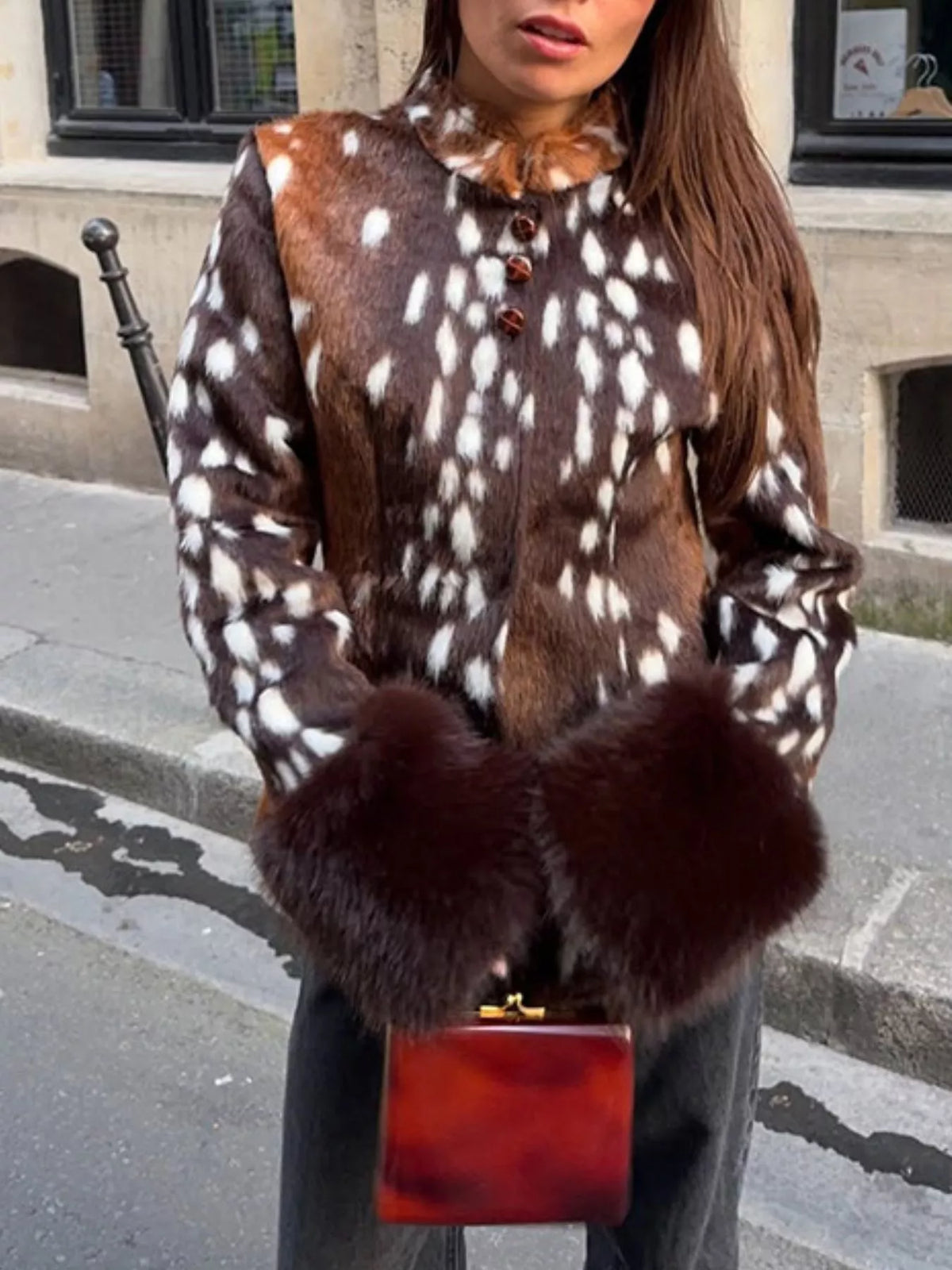 A woman wearing a brown and white spotted faux fur jacket with dark brown fur cuffs, holding a small tortoiseshell clutch bag, standing on a city street.