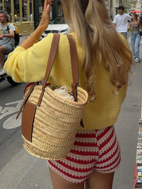 Woman in yellow sweater and red and white shorts carrying a straw bag on a city street.