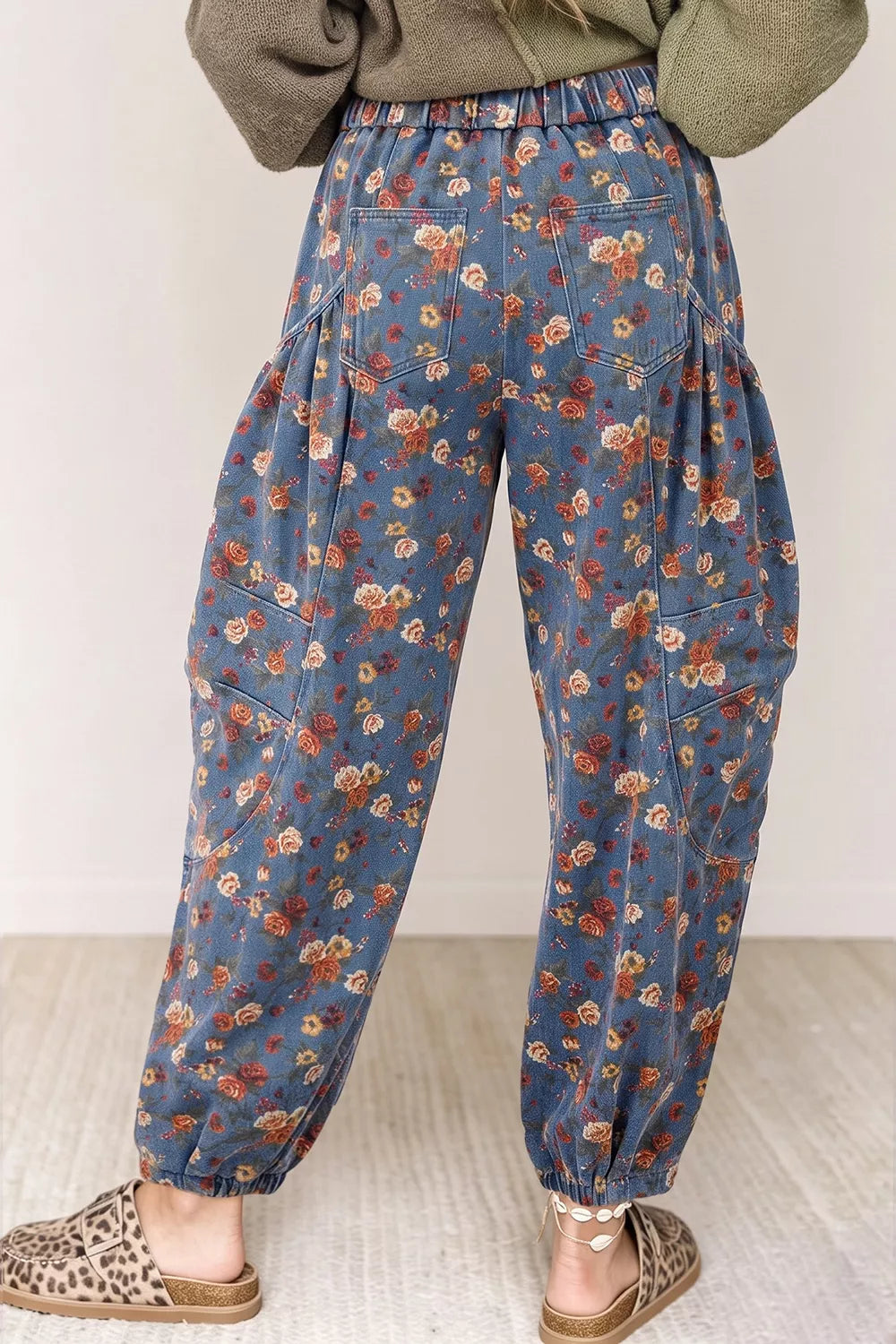 A person shown from the back wearing loose, baggy blue floral pants with an elastic waist and ankle cuffs, paired with leopard-print slippers and an olive green top.
