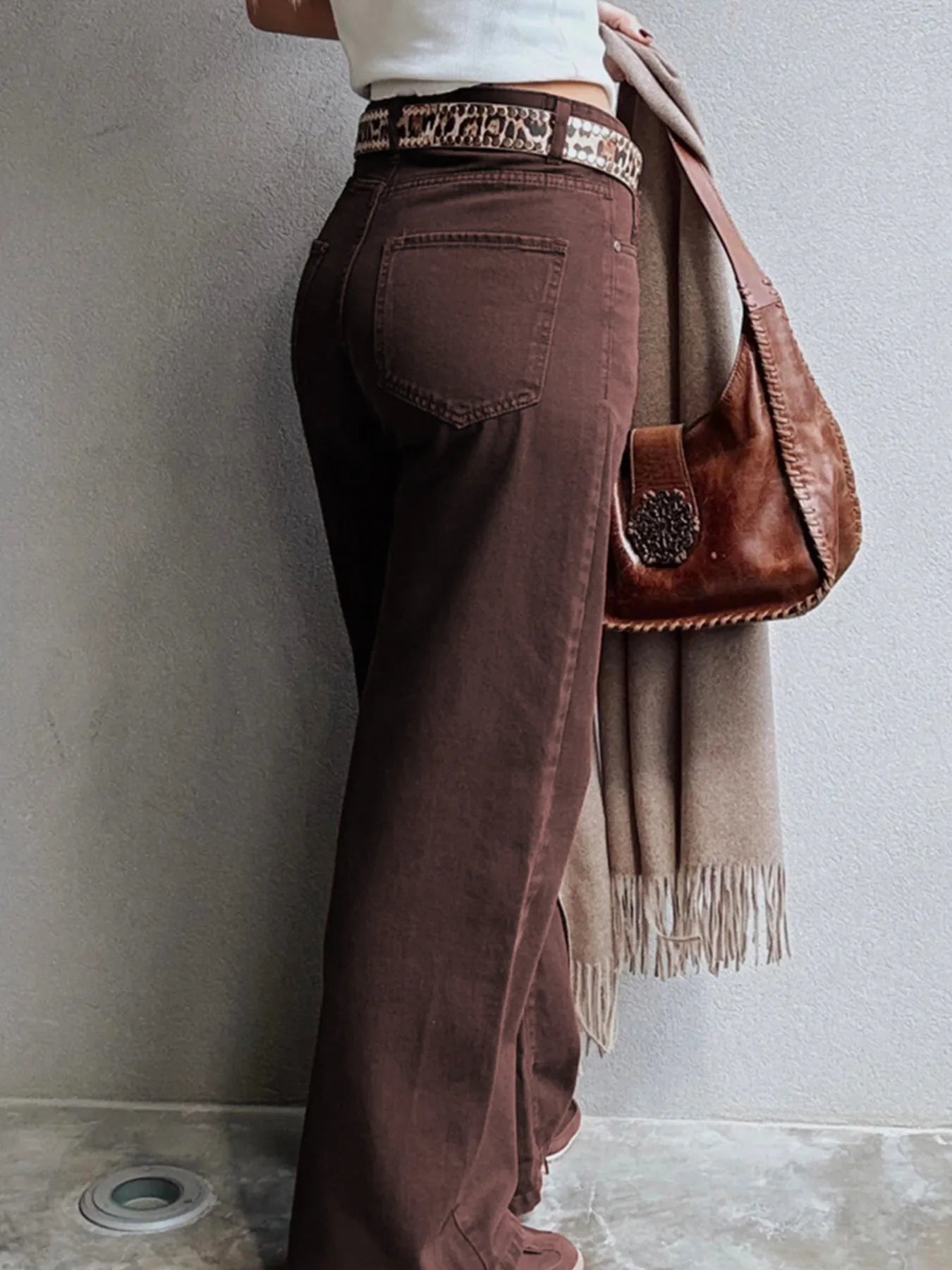 Person wearing brown pants with a patterned belt, holding a brown bag and beige scarf against a gray wall.