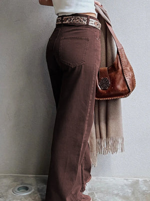Person wearing brown pants with a patterned belt, holding a brown bag and beige scarf against a gray wall.