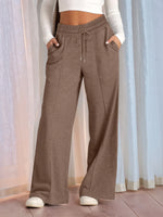 Brown wide-leg pants worn by a person with a white top and shoes, against a light curtain background.