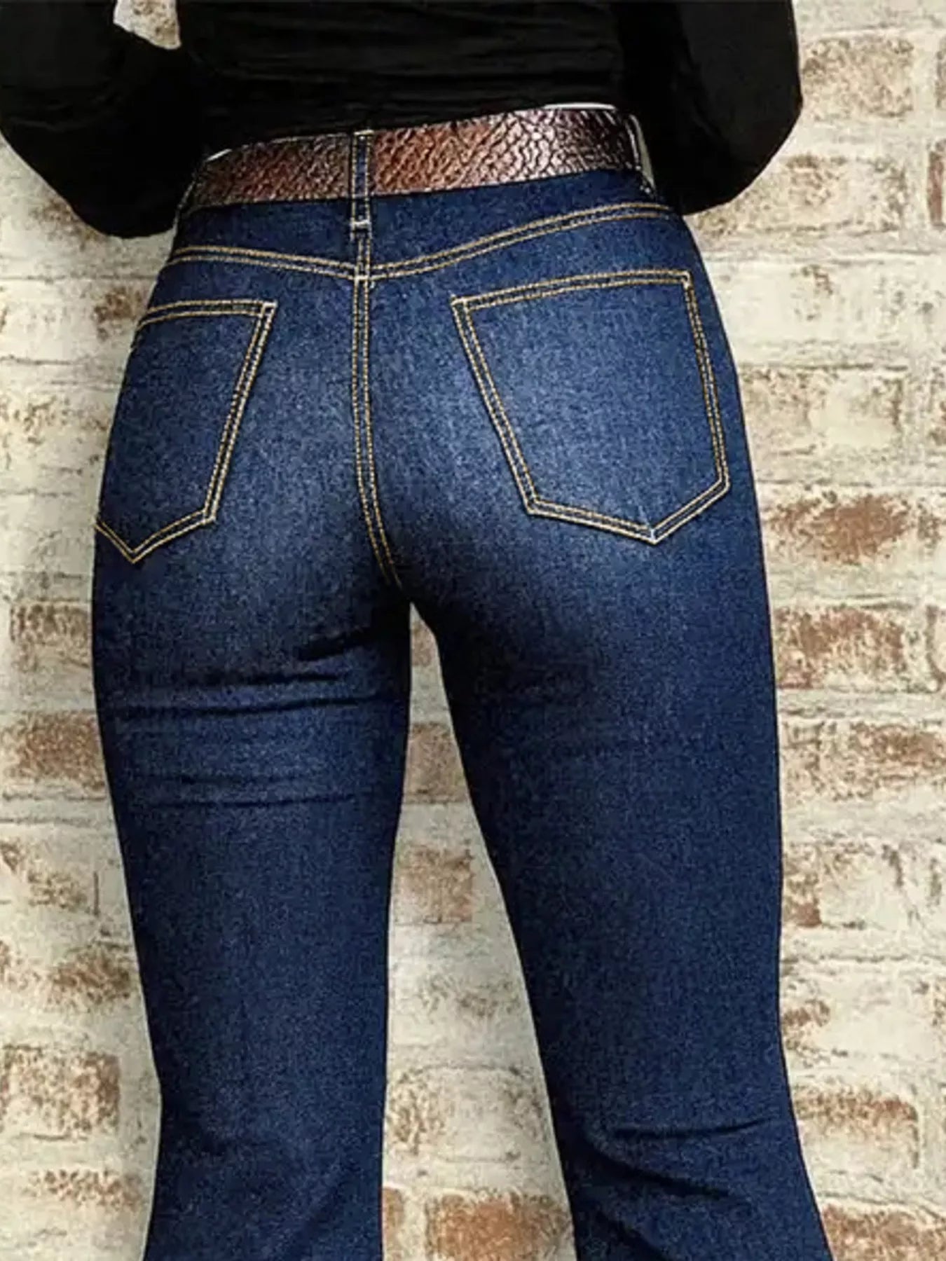 Person wearing blue jeans with a brown belt against a brick wall.