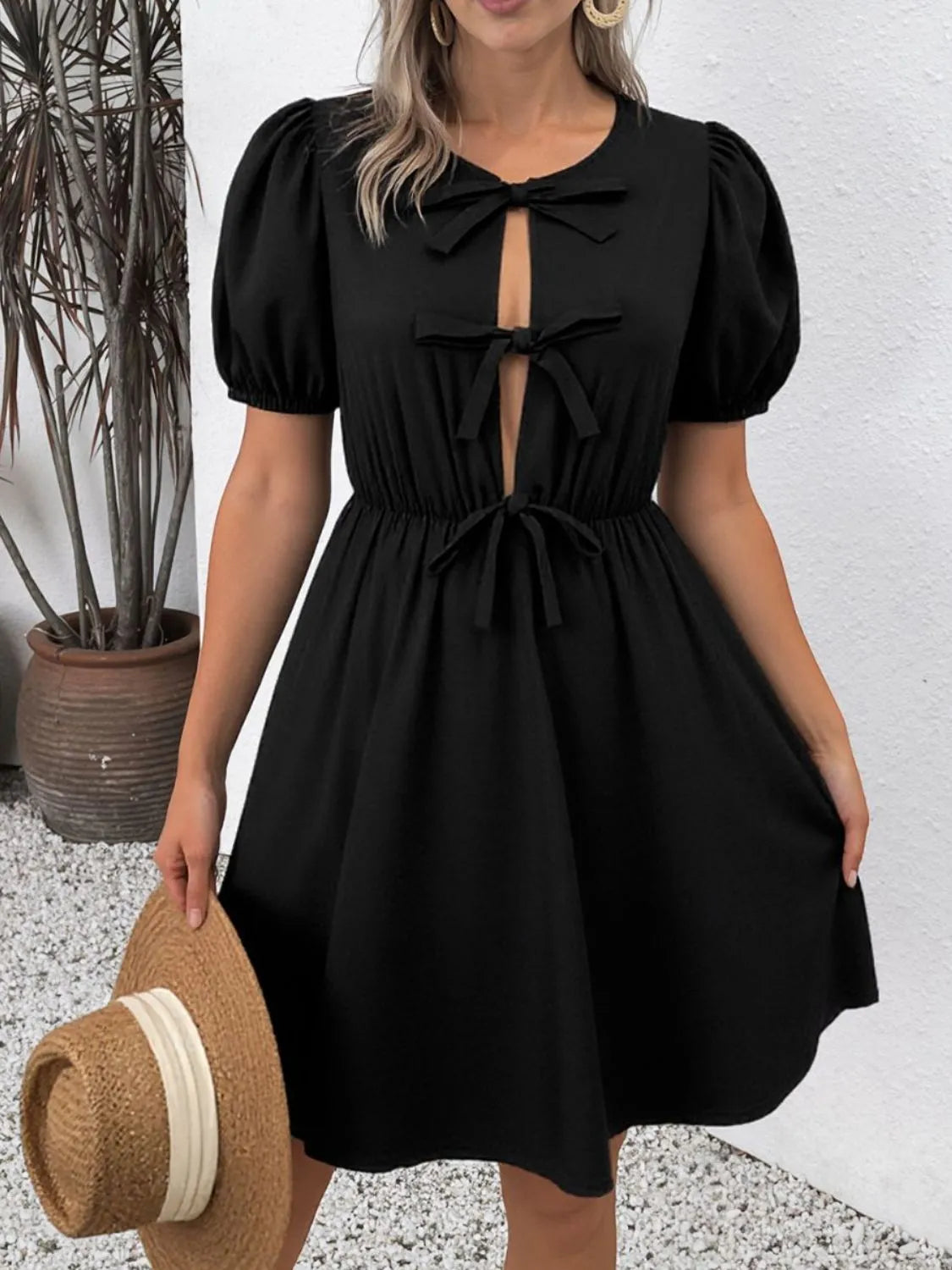 Woman wearing a black dress with puff sleeves holding a straw hat against a white wall.