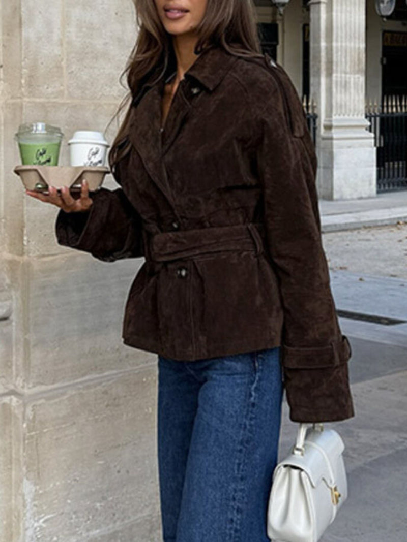 Person wearing a brown suede jacket holding a white handbag and a green drink.