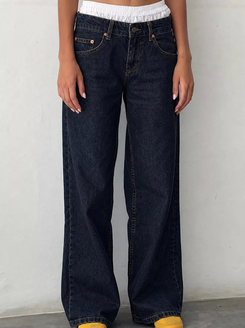 A person wearing dark indigo wide-leg jeans with a white cropped top and yellow shoes, standing against a plain light gray wall.