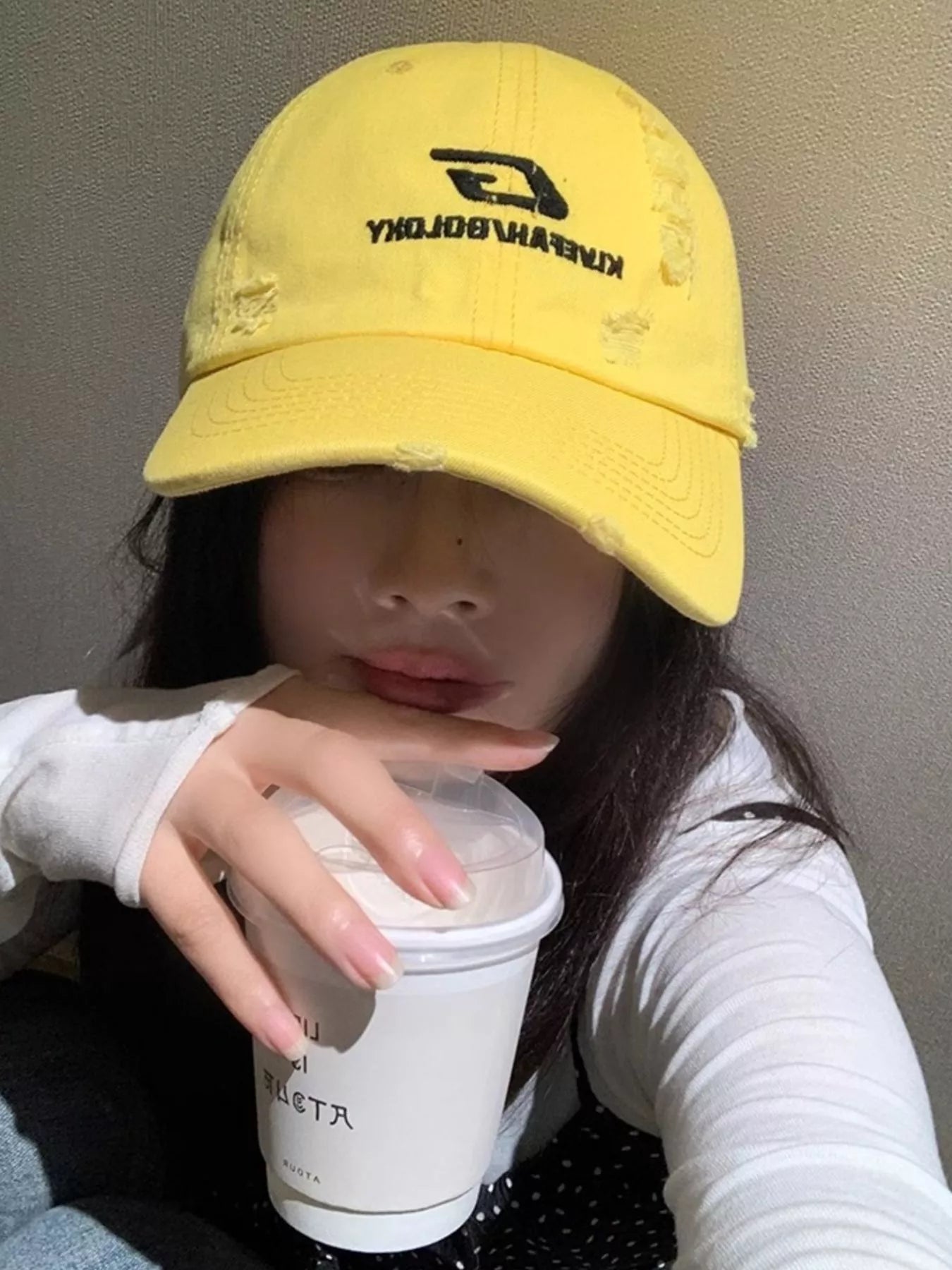Person wearing a yellow cap with a brand logo, holding a white cup.