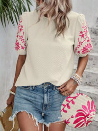 Woman wearing a beige top with pink floral embroidery and denim shorts, holding a pink and white bag.