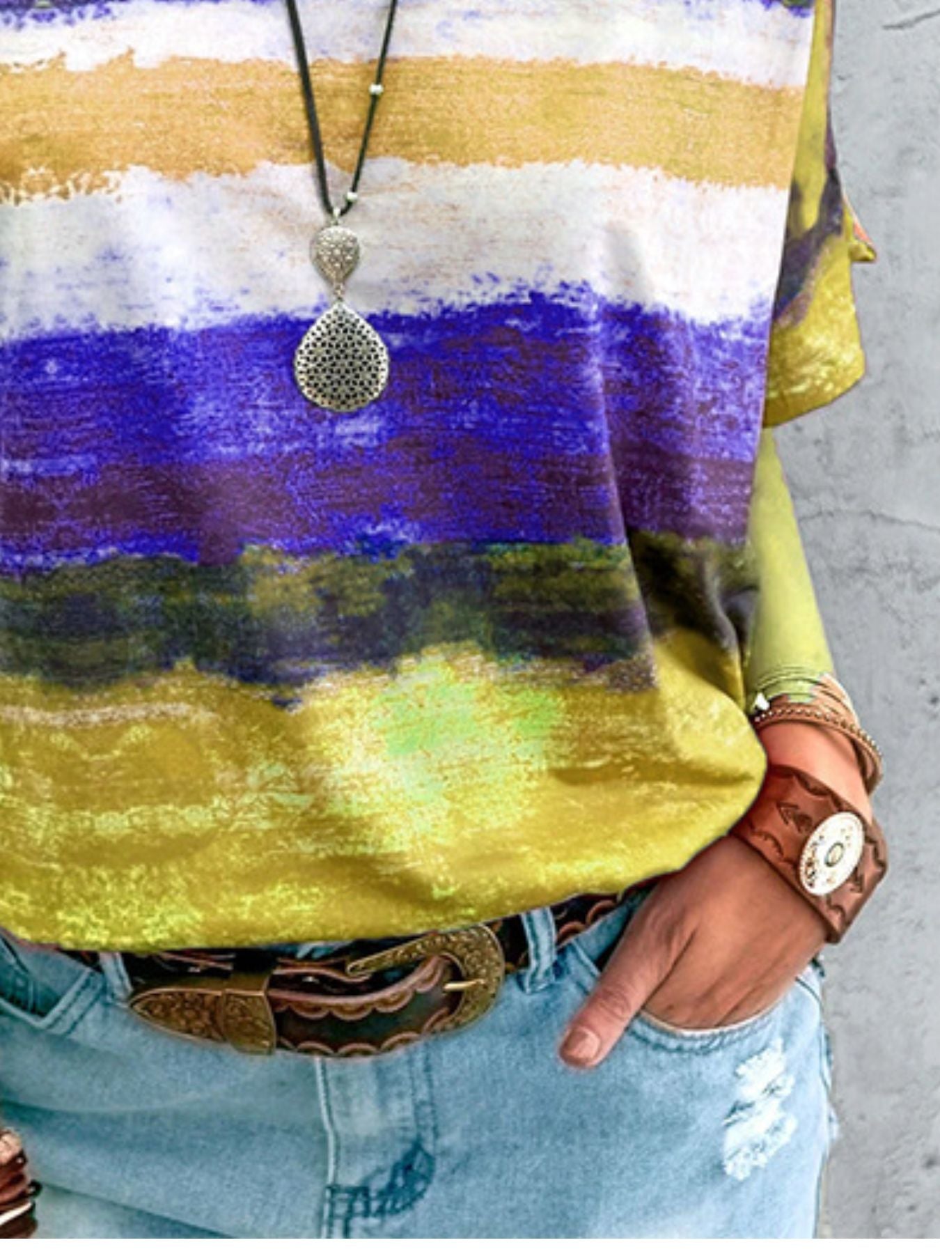 Person wearing a colorful striped shirt with a necklace and denim jeans on a concrete surface.