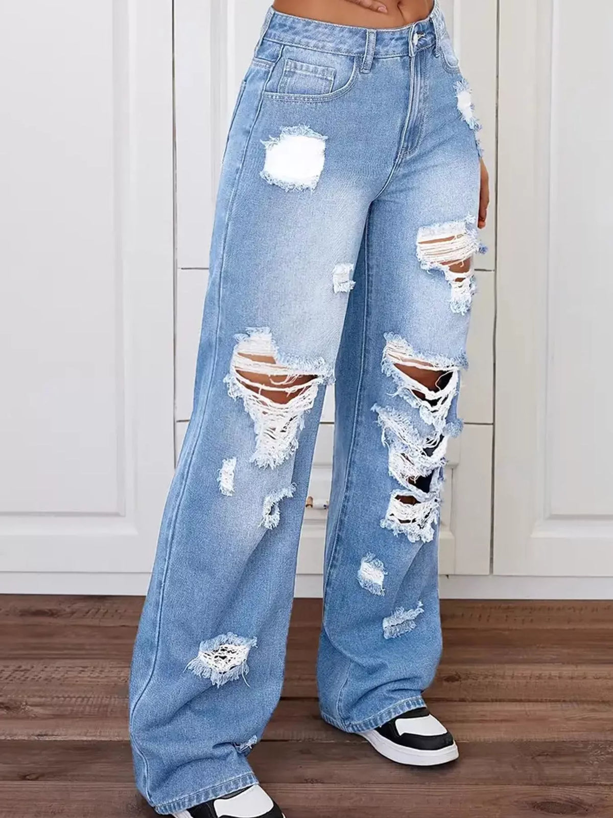 Light blue wide-leg denim jeans with heavy distressing, rips, and frayed details, shown on a model wearing black and white sneakers against a white paneled wall and wood floor.