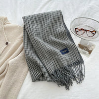 Gray checkered scarf with a brand logo on a white surface next to a beige cardigan and glasses.