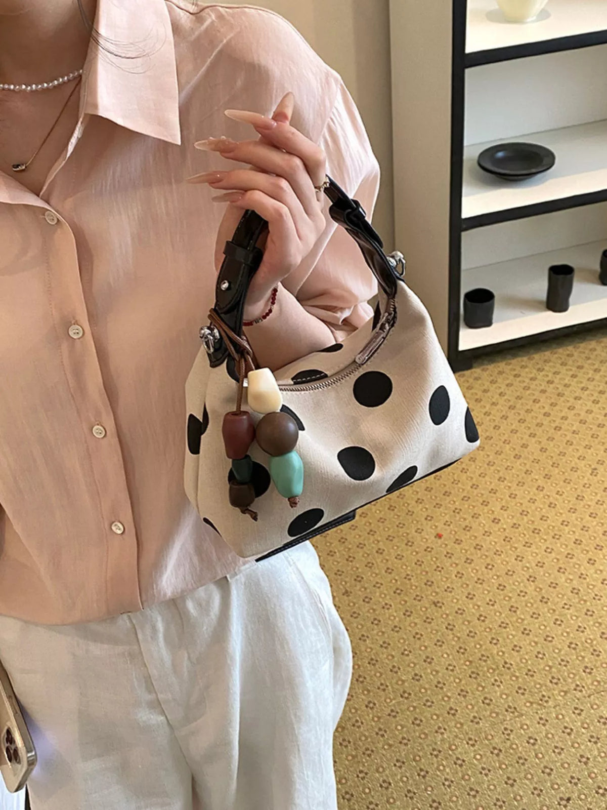A person wearing a pale pink button-up shirt and white pants holds a small shoulder bag with a white base and large black polka dots. The bag has dark brown trim, a top handle, and a decorative cluster of wooden beads hanging from the strap.